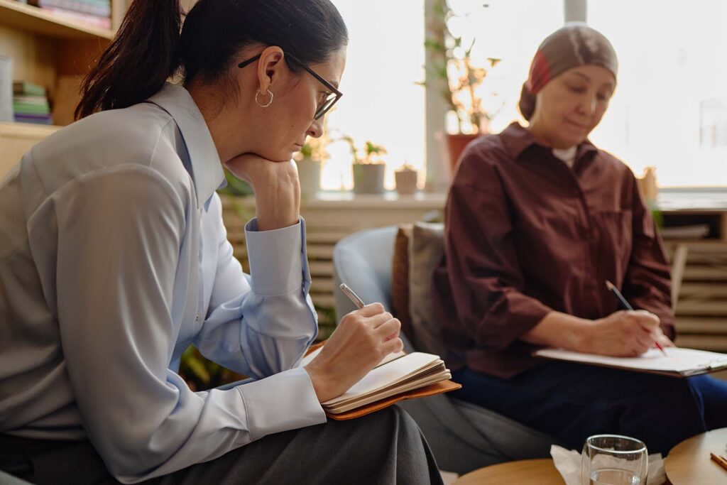 Middle aged Caucasian woman with ponytail sitting and writing in notebook while observing senior woman with headscarf completing psychological assessment in office setting