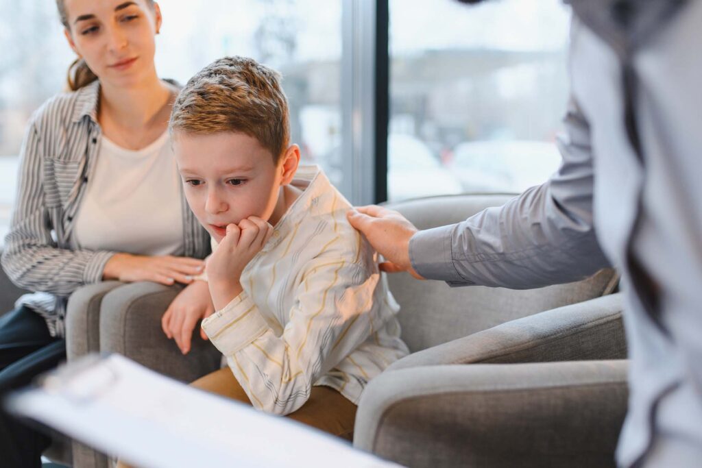 Child feeling anxious or shy, receiving comfort from a psychologist. Mother observing during a family counseling session