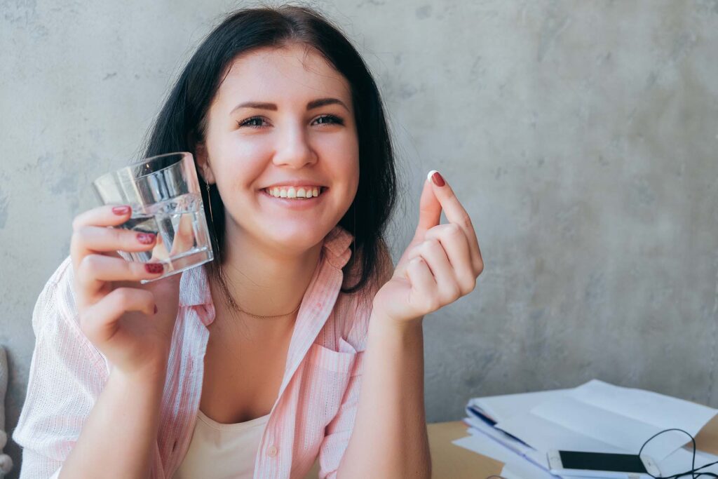 A beautiful girl smiling, not a healthy business woman holding a pill and a glass of water. A female teenager is drinking medicine, an antidepressant and is happy