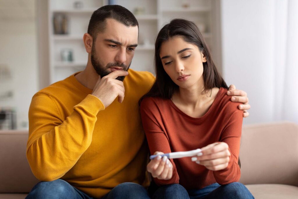 Infertility Problems. Portrait Of Upset Young Couple Looking At Negative Pregnancy Test While Sitting Together On Couch At Home, Caring Husband Embracing Depressed Wife, Free Space